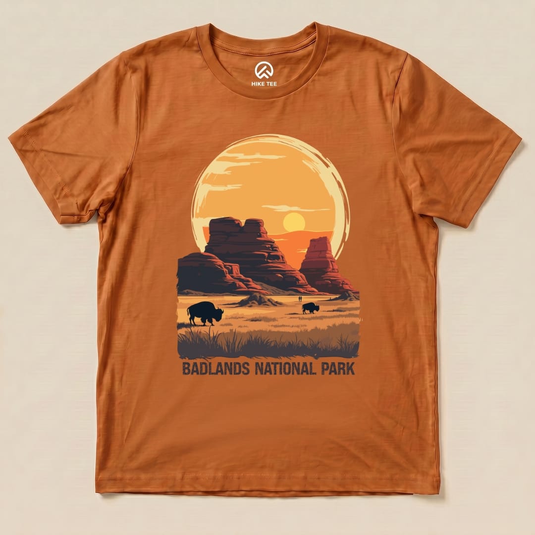A flat graphic t-shirt in burnt orange featuring a sunset over Badlands National Park with bison and "BADLANDS NATIONAL PARK" text.