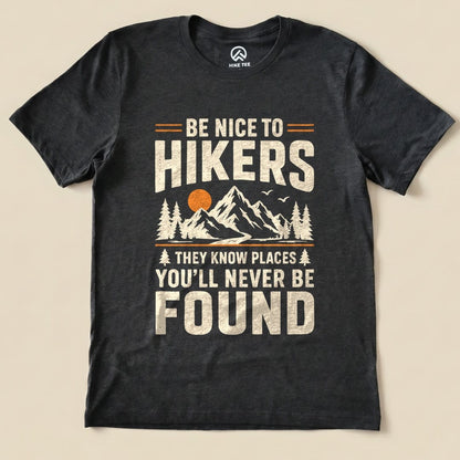 A graphic t-shirt featuring the phrase "Be Nice To Hikers" along with mountains, trees, and a sun design in earthy colors.