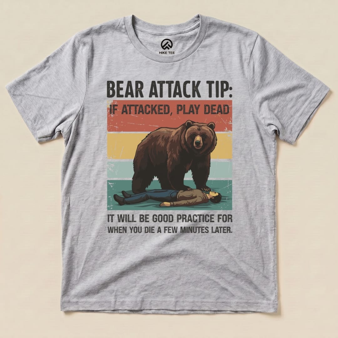 A flat graphic t-shirt features a bear and a collapsed person, with the text “Bear Attack Tip: If attacked, play dead.”