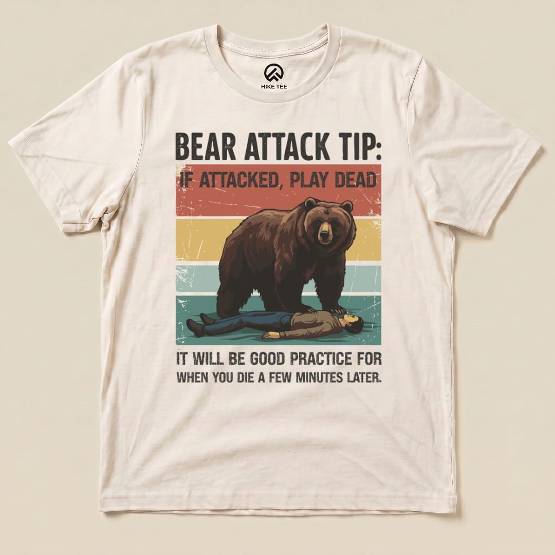 A graphic t-shirt featuring a bear looming over a person lying on the ground with the text "Bear Attack Tip: If attacked, play dead" and additional wording underneath.