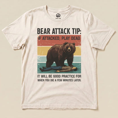 A graphic t-shirt featuring a bear looming over a person lying on the ground with the text "Bear Attack Tip: If attacked, play dead" and additional wording underneath.
