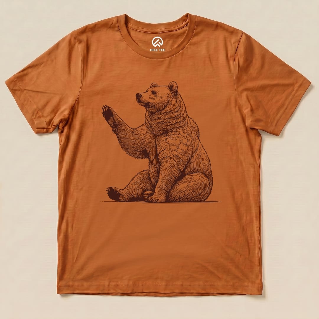 A graphic t-shirt in rust color featuring a whimsical depiction of a bear waving, labeled as the "Bear is Watching Shirt.