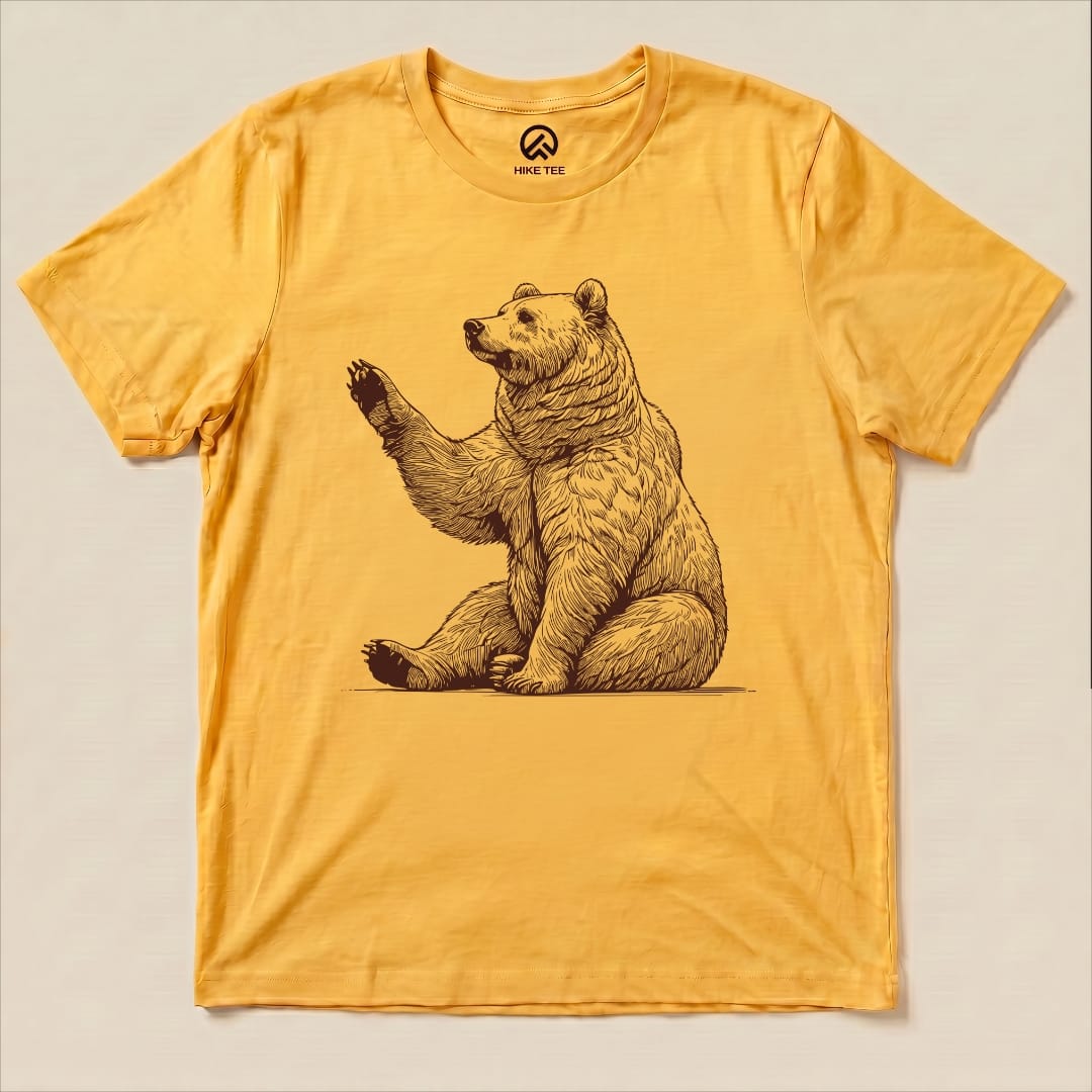 A graphic t-shirt in mustard yellow featuring a waving bear illustration, labeled the "Bear is Watching Shirt.