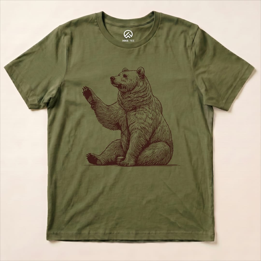 The olive green graphic t-shirt features a bear waving, with the design capturing a playful essence, labeled "Bear is Watching Shirt.