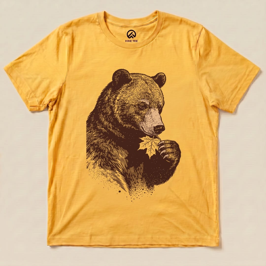 A flat lay of the Bear With Leaf Shirt features a bear holding a leaf printed in dark brown on a vibrant yellow graphic t-shirt.
