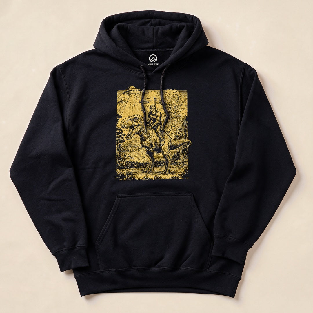 The Bigfoot Dinosaur Hoodie features a whimsical graphic of Bigfoot riding a dinosaur in a forested landscape.