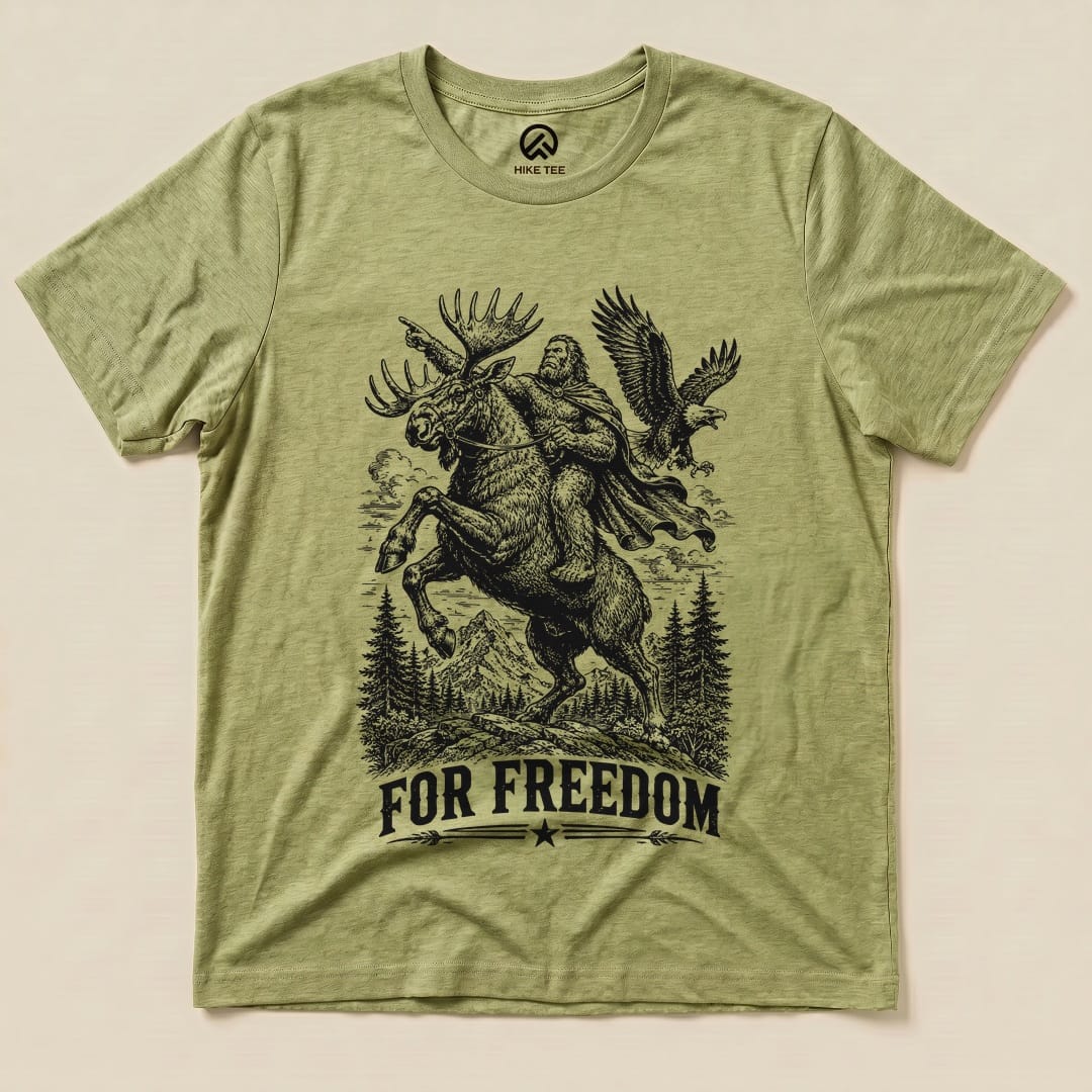 A graphic t-shirt featuring a whimsical design of Bigfoot riding a moose with an eagle overhead and the phrase "FOR FREEDOM.