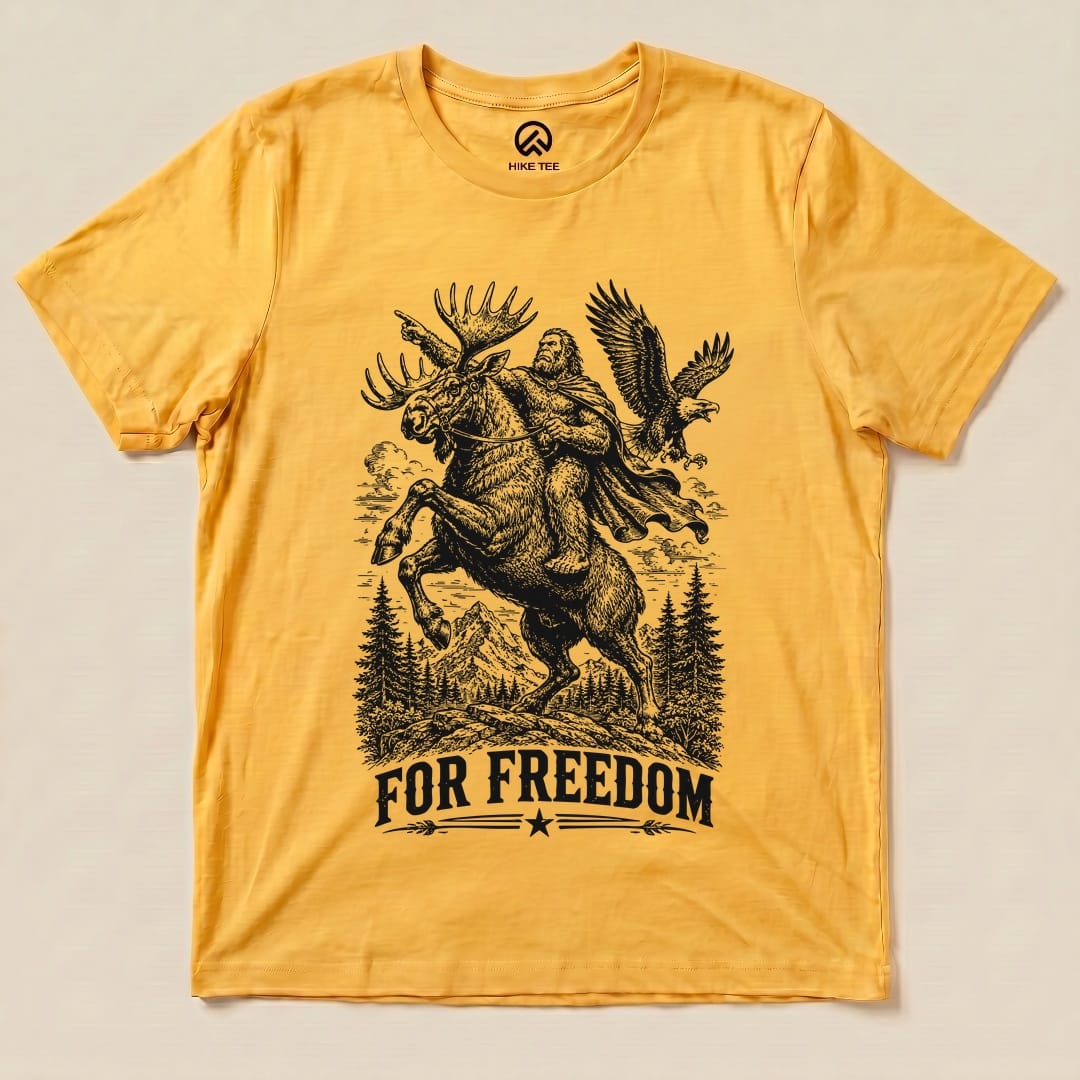 A graphic t-shirt featuring a whimsical illustration of Bigfoot riding a moose, accompanied by an eagle, with the text "FOR FREEDOM" below.