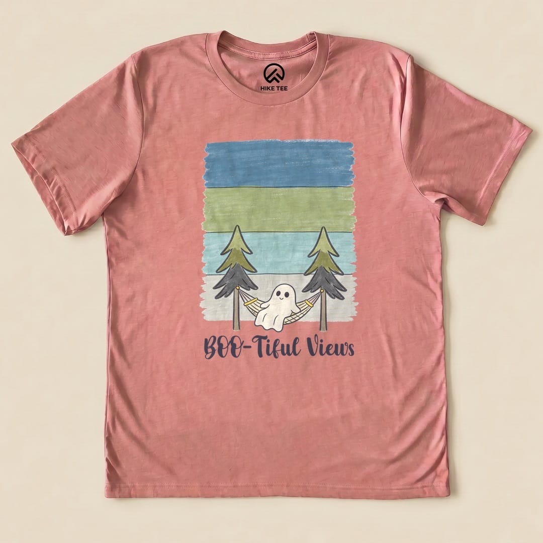 A graphic t-shirt in soft pink featuring a playful ghost hanging in a hammock between pine trees with the text "Boo-tiful Views.