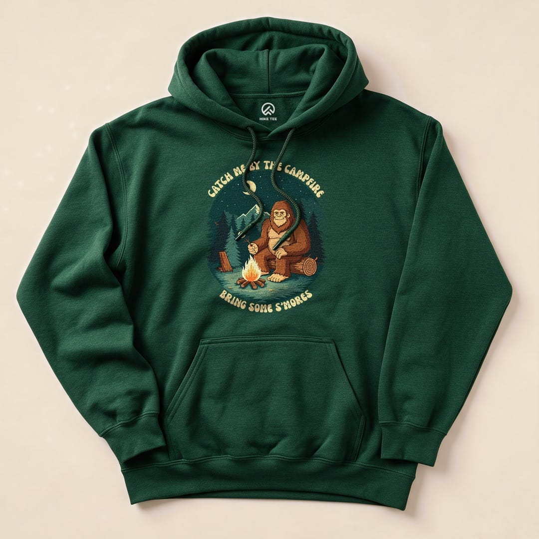 A green hoodie featuring a graphic of a Sasquatch by a campfire with the text "Catch Me By The Campfire, Bring Some S'mores".