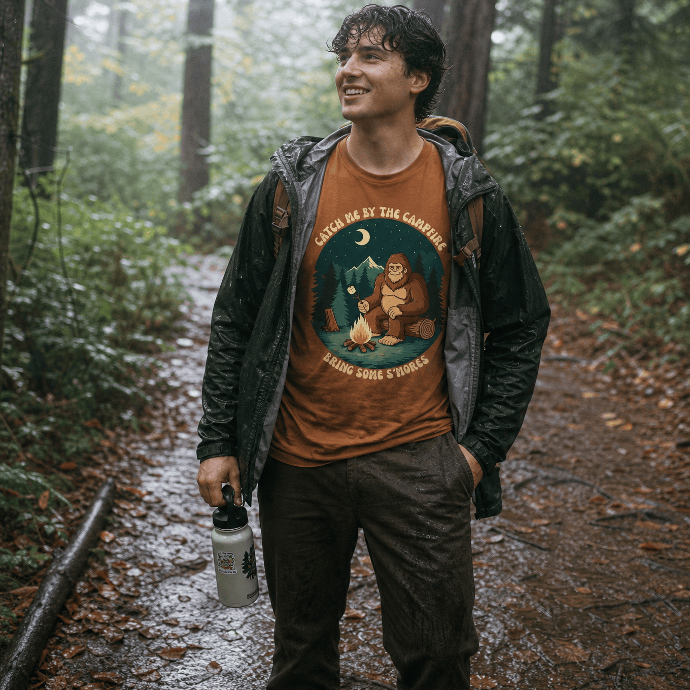 A man stands in a rainy forest wearing a brown graphic t-shirt featuring a bear by a campfire with the text “Bring Some S'mores Shirt.”
