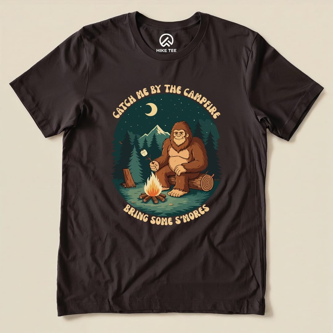 A graphic t-shirt featuring a cartoon Sasquatch by a campfire with the phrases "Catch Me by the Campfire" and "Bring Some S'mores.