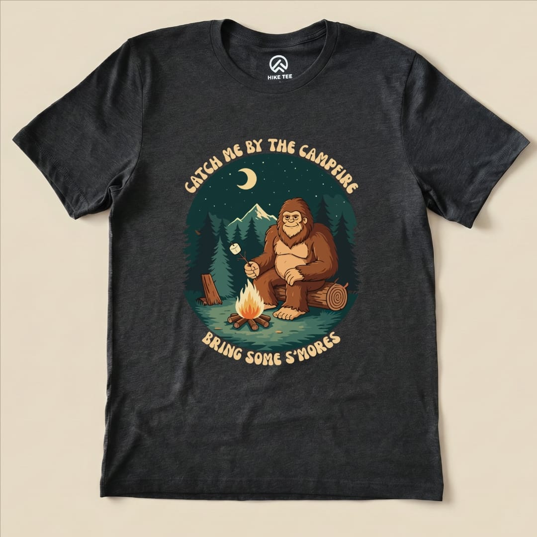 Graphic t-shirt featuring a Sasquatch sitting by a campfire, with the slogan "Bring Some S'mores" and surrounding trees and mountains.