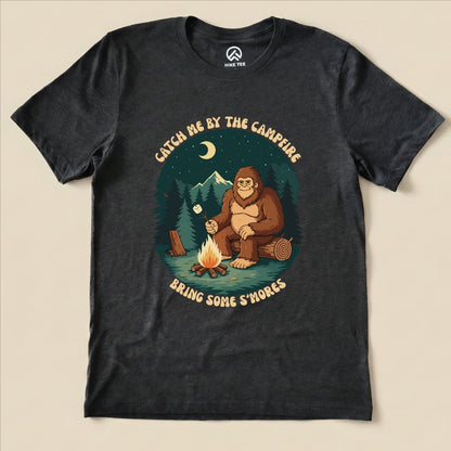 Graphic t-shirt featuring a Sasquatch sitting by a campfire, with the slogan "Bring Some S'mores" and surrounding trees and mountains.