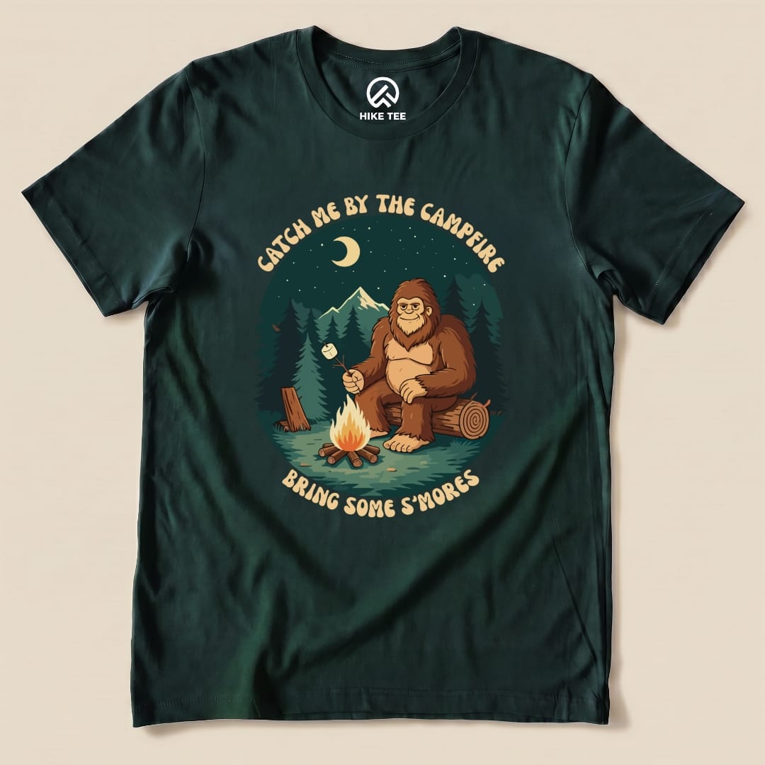 Graphic t-shirt featuring a cartoon bigfoot by a campfire with the slogan "Catch Me by the Campfire" and "Bring Some S'mores.
