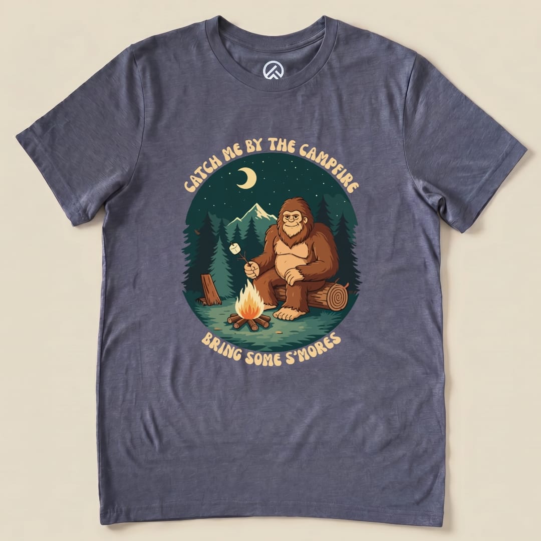 Graphic t-shirt featuring a humorous design of a bigfoot by a campfire with the slogan "Catch me by the campfire, Bring Some S'mores.