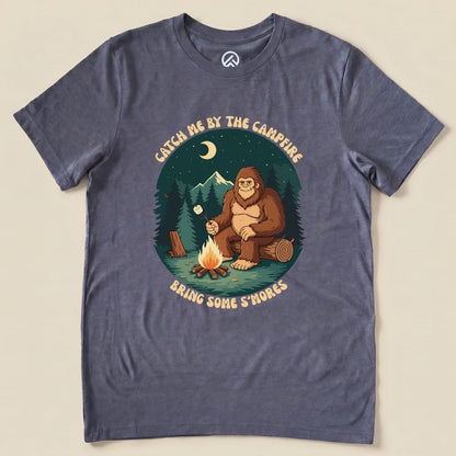 Graphic t-shirt featuring a humorous design of a bigfoot by a campfire with the slogan "Catch me by the campfire, Bring Some S'mores.