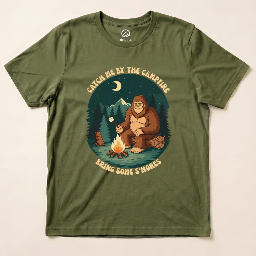 The olive green graphic t-shirt features a cartoon Bigfoot by a campfire, with the slogan "Bring Some S'mores" above.