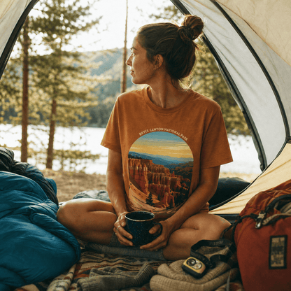 A woman sits inside a tent, holding a cup, wearing a graphic t-shirt featuring the "Bryce Canyon National Park Shirt" design.