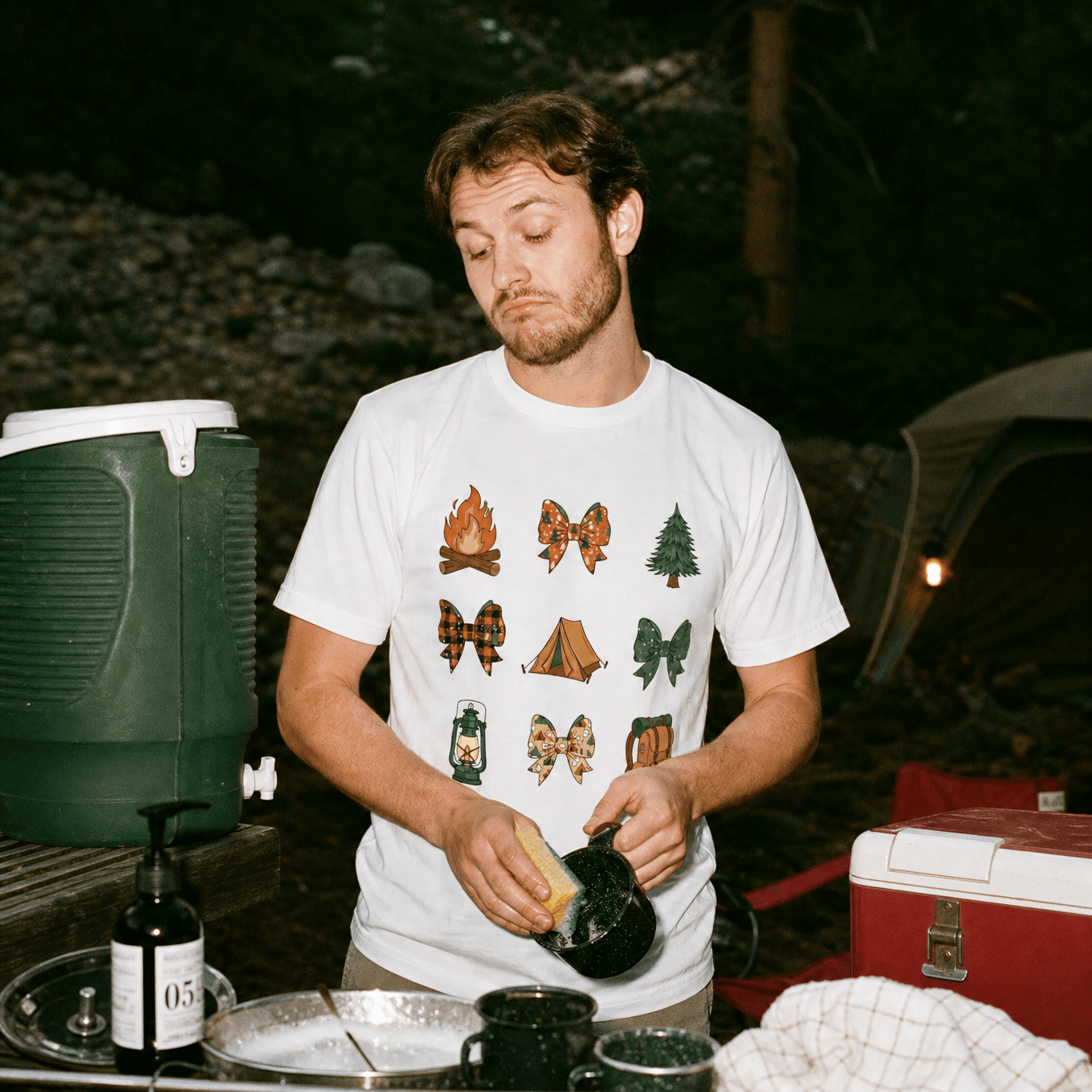 A man stands at a camping setup wearing a graphic t-shirt featuring various camping bows and icons, labeled as "Camping Bows Shirt.