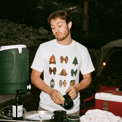 A man stands at a camping setup wearing a graphic t-shirt featuring various camping bows and icons, labeled as "Camping Bows Shirt.