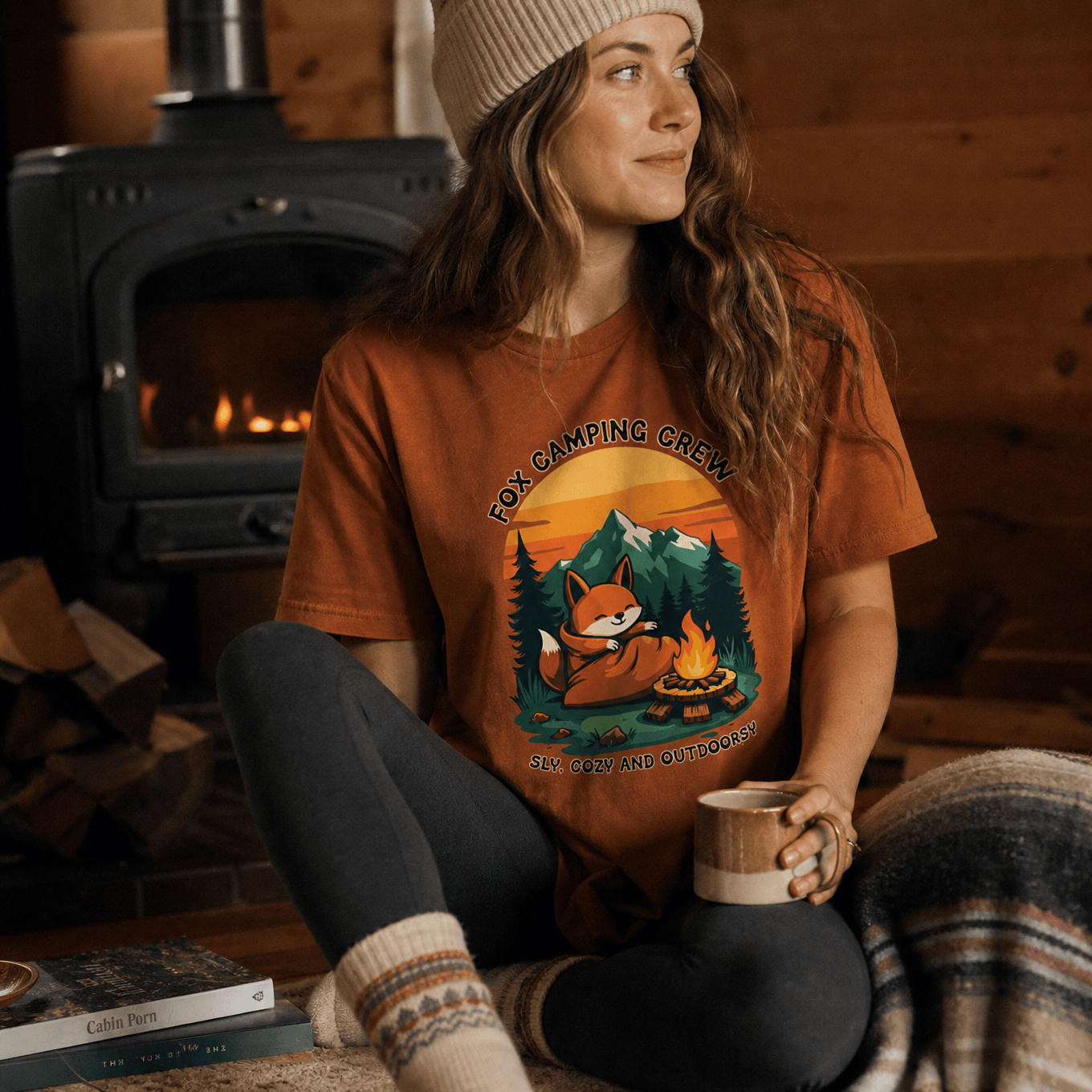 A woman sits comfortably in a cabin, wearing a Camping Fox Shirt featuring a relaxed fox by a campfire with the text "Fox Camping Crew.