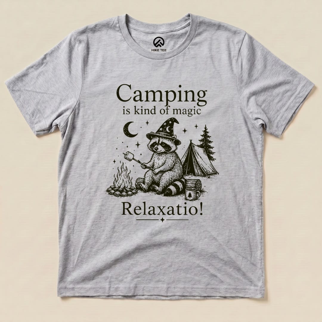 A graphic t-shirt featuring a raccoon wearing a wizard hat by a campfire, with the phrase "Camping is kind of magic" above.