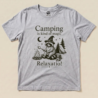 A graphic t-shirt featuring a raccoon wearing a wizard hat by a campfire, with the phrase "Camping is kind of magic" above.