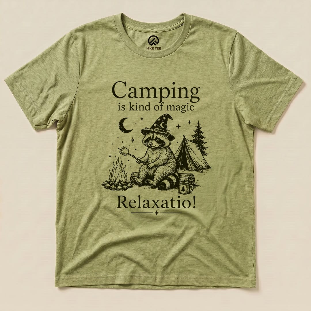 A graphic t-shirt in olive green featuring a raccoon with a wizard hat and a campfire, stating "Camping is kind of magic Relaxatio!