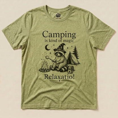 A graphic t-shirt in olive green featuring a raccoon with a wizard hat and a campfire, stating "Camping is kind of magic Relaxatio!