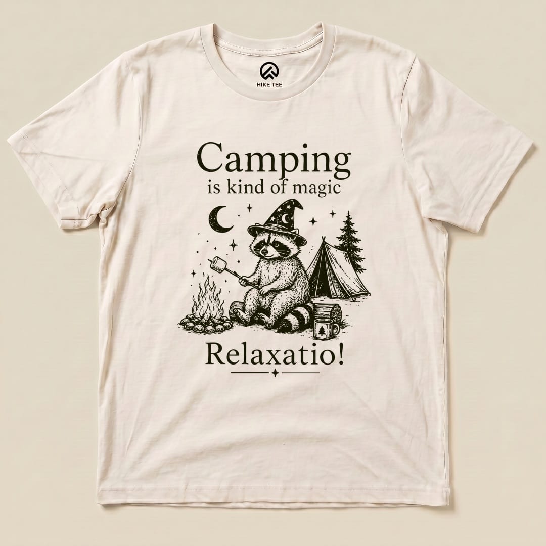 A flat graphic t-shirt featuring a raccoon in a wizard hat, with the slogan "Camping is kind of magic Relaxatio!" and a camping scene.