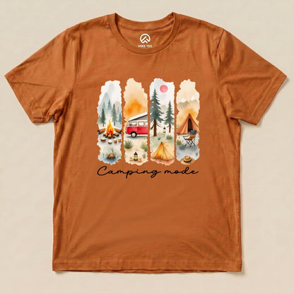 A graphic t-shirt in a warm orange color featuring a colorful camping scene including a retro camper, trees, and the phrase "Camping mode.