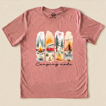 A pink graphic t-shirt featuring a watercolor design with camping scenes and the phrase "Camping mode" printed at the bottom.