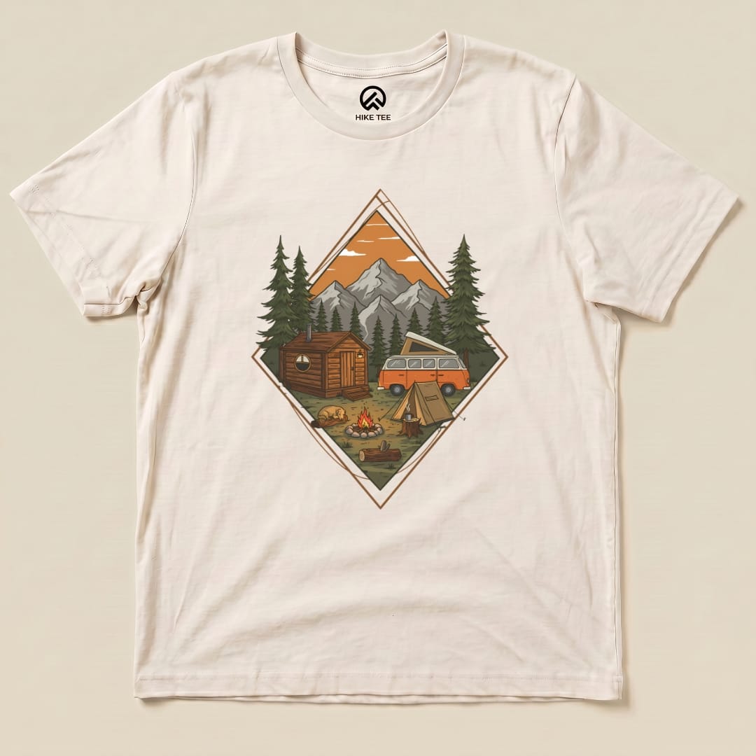 A graphic t-shirt featuring a detailed camping scenery design with a cabin, camper van, trees, and a campfire.