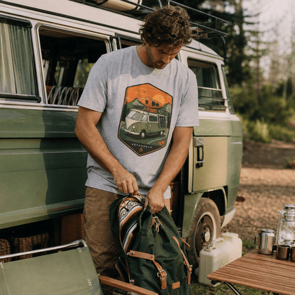 A man wearing a graphic t-shirt with a "Camping Van Adventure Shirt" design featuring a vintage camper and mountains, prepares his backpack outdoors.