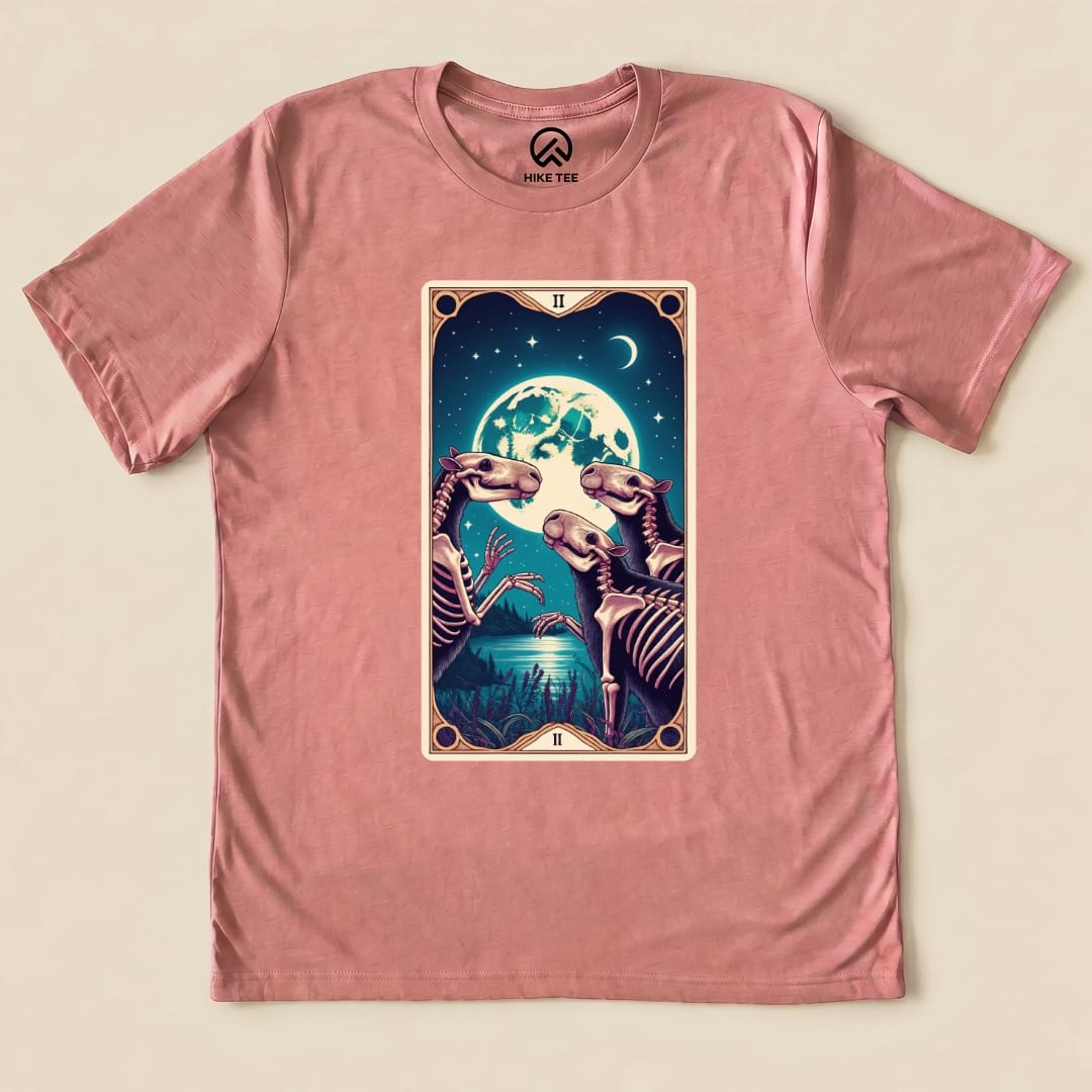 A graphic t-shirt in pink featuring a Capybara Moon Tarot Card design with skeletons beneath a large moon and stars.