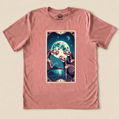 A graphic t-shirt in pink featuring a Capybara Moon Tarot Card design with skeletons beneath a large moon and stars.