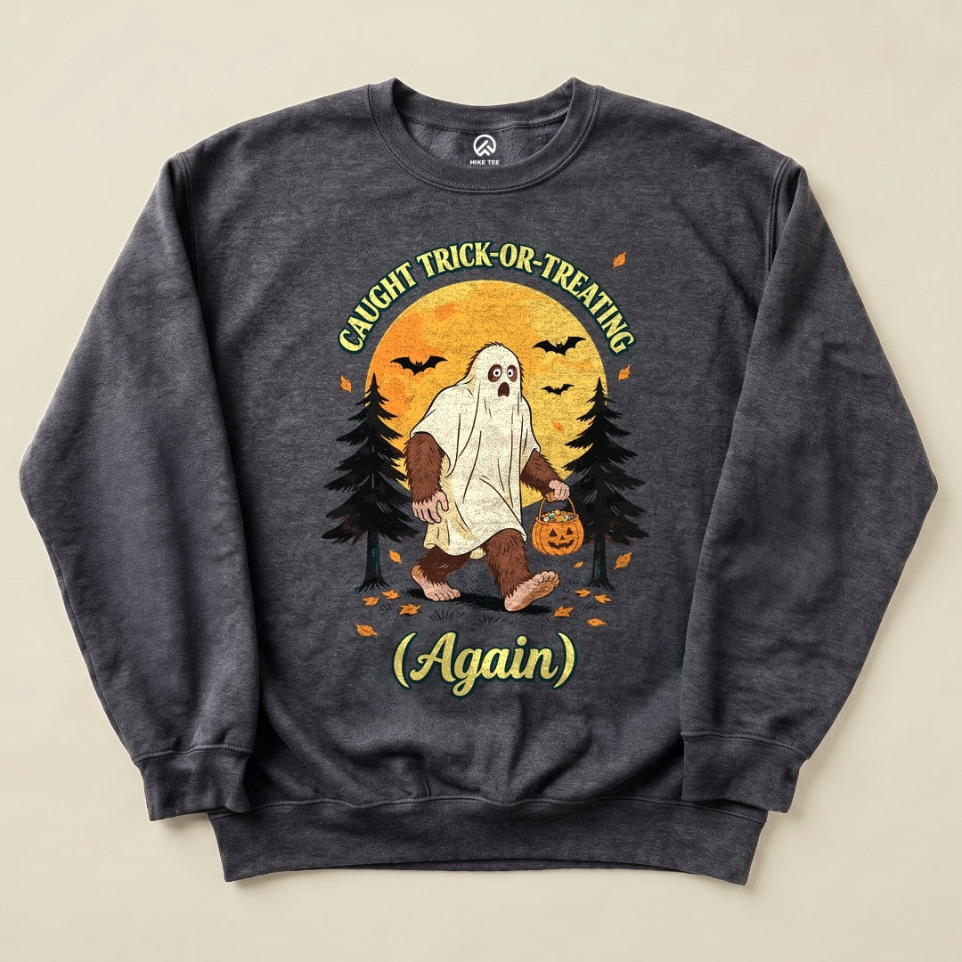 A graphic sweatshirt featuring a bigfoot in a ghost costume holding a pumpkin, with the words "Caught Trick-or-Treating Again.