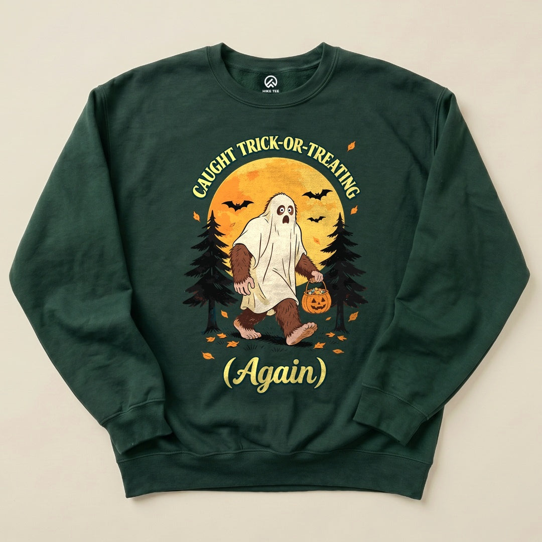 Caught Trick-or-Treating Again Sweatshirt featuring a bigfoot in a ghost costume with a pumpkin, surrounded by trees and bats.