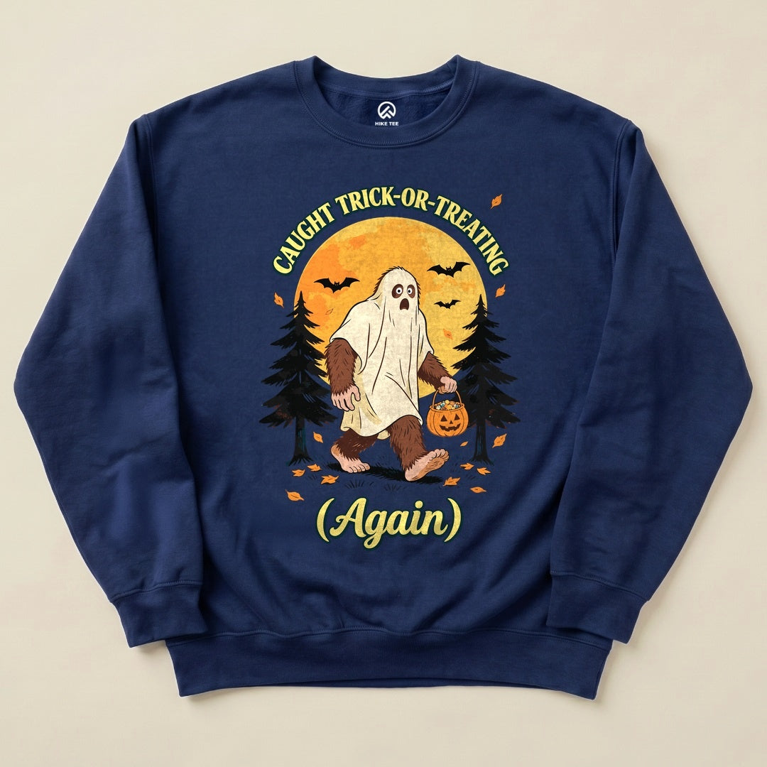 A graphic sweatshirt featuring a Bigfoot in a ghost costume carrying a pumpkin, with the phrase "Caught Trick-or-Treating Again.