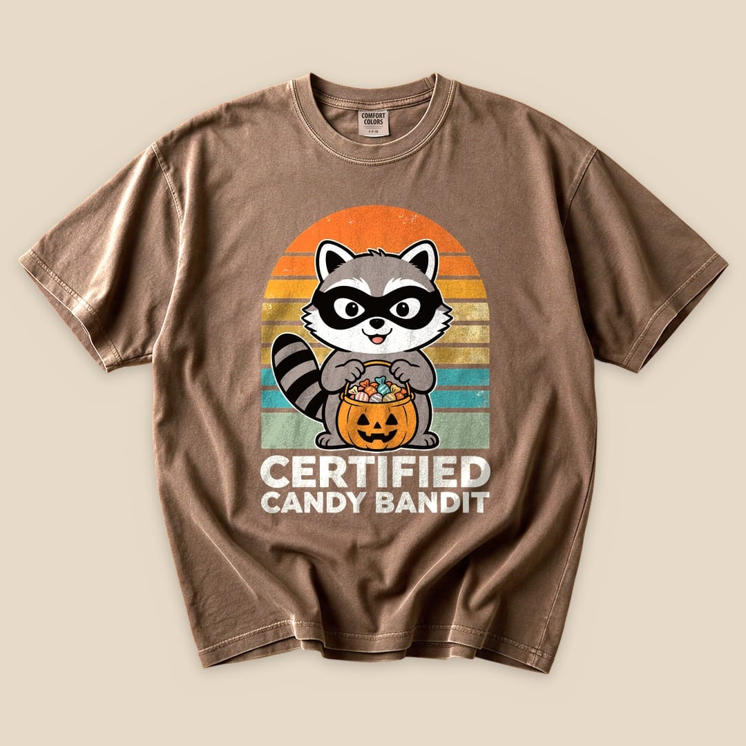 A brown graphic t-shirt featuring a playful raccoon holding a pumpkin full of candy, labeled "Certified Candy Bandit.