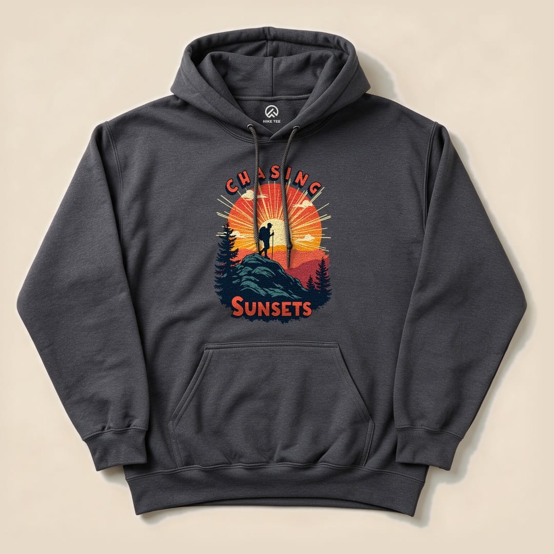 A dark gray Chasing Sunsets Hoodie features a graphic of a hiker on a rock against a vibrant sunset with trees.