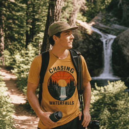 A man stands in a forest wearing a graphic t-shirt with a colorful design that reads "Chasing Waterfalls Retro Shirt" above a waterfall.