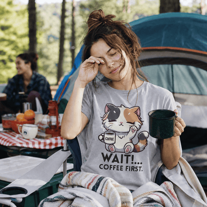 A woman sitting in a camping chair holds a mug, wearing a graphic t-shirt featuring a cat and the text "WAIT!... COFFEE FIRST.