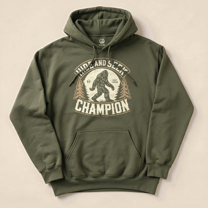 The olive green Hide and Seek Champion Bigfoot Hoodie features a graphic of Bigfoot with trees and bold text in a vintage style.