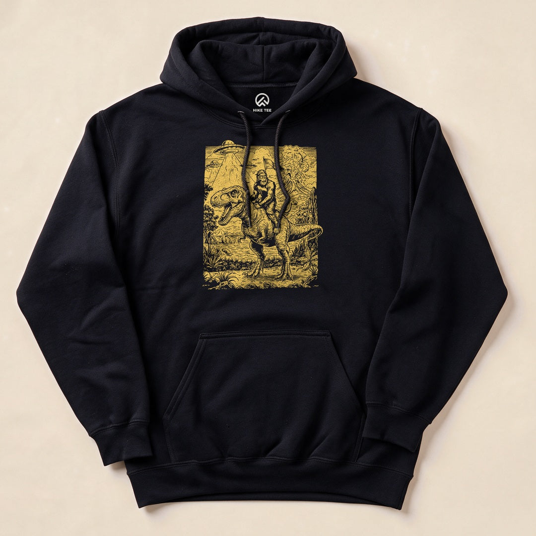The Bigfoot Dinosaur Hoodie features a whimsical graphic of Bigfoot riding a dinosaur in a forested landscape.