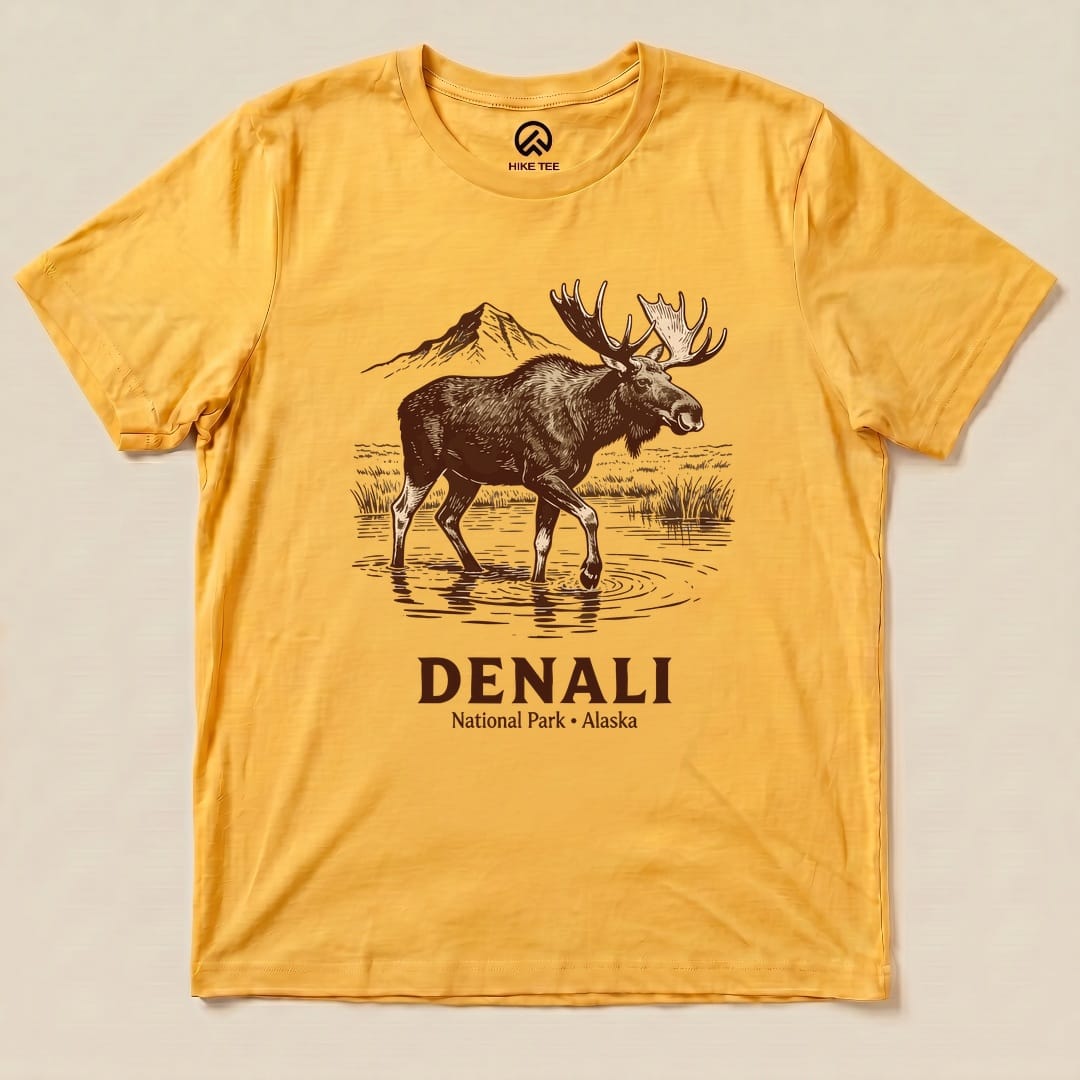 A graphic t-shirt in yellow featuring a moose standing in water with mountains, labeled "Denali National Park - Alaska.