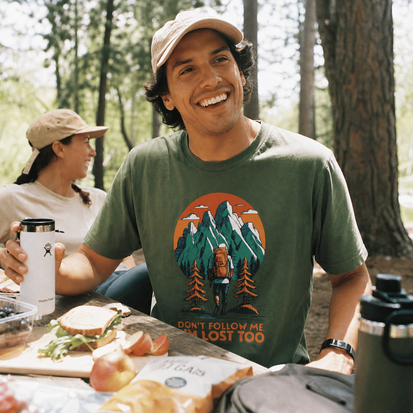 Smiling man at a picnic table in the woods wears a "Don't Follow Me Shirt," featuring a hiker graphic with mountains.