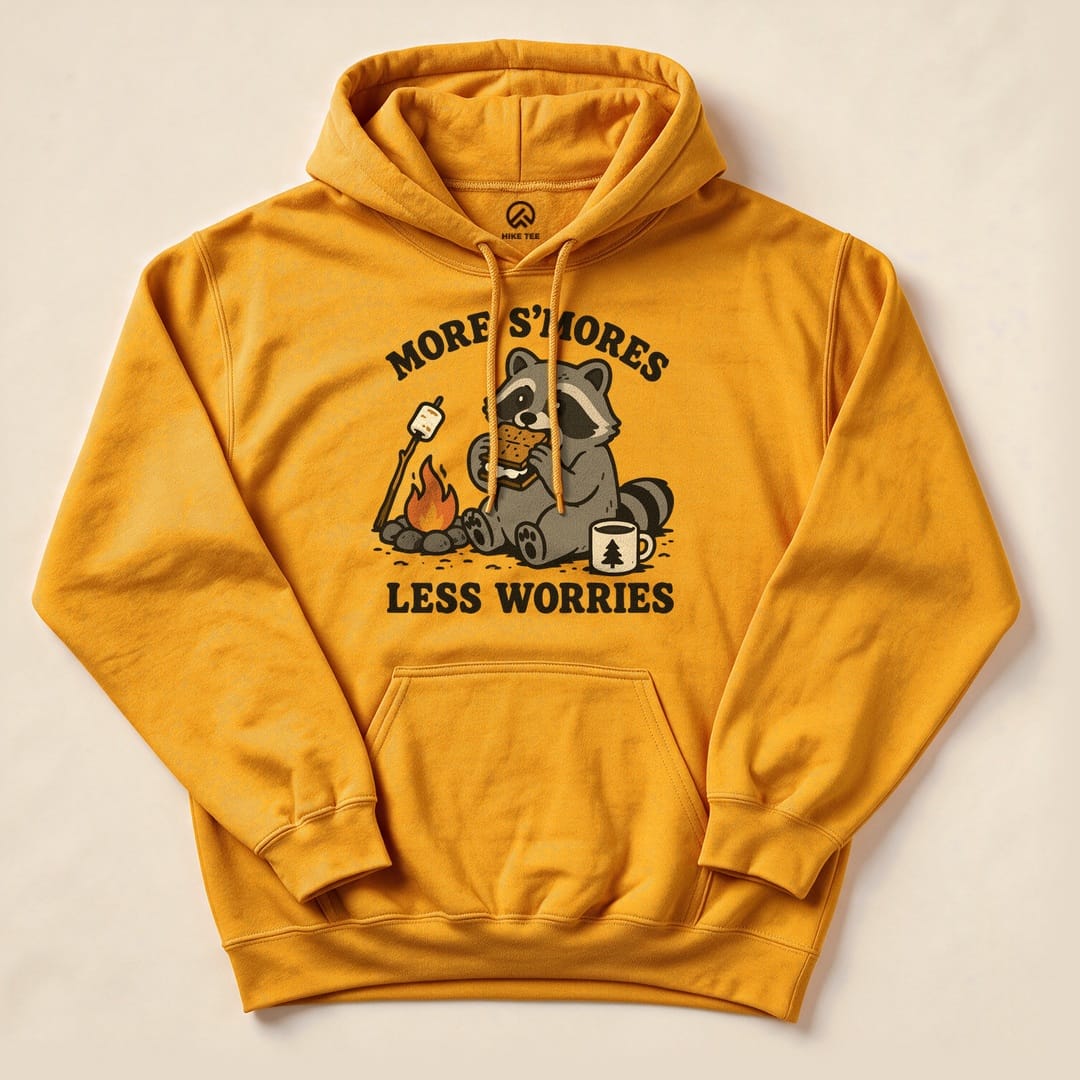A graphic hoodie in mustard yellow featuring a raccoon enjoying s'mores by a campfire, with the text "More S'mores Less Worries.