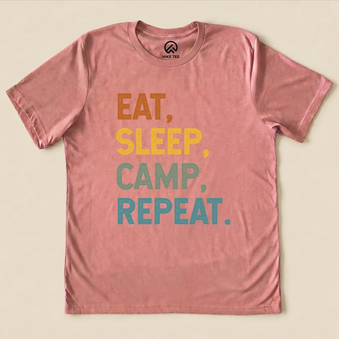 A flat graphic t-shirt in a soft pink hue features the slogan "Eat Sleep Camp Repeat" in playful, colorful lettering.